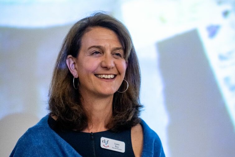 Woman smiling in front of a presentation screen, wearing a name tag and blue shawl.