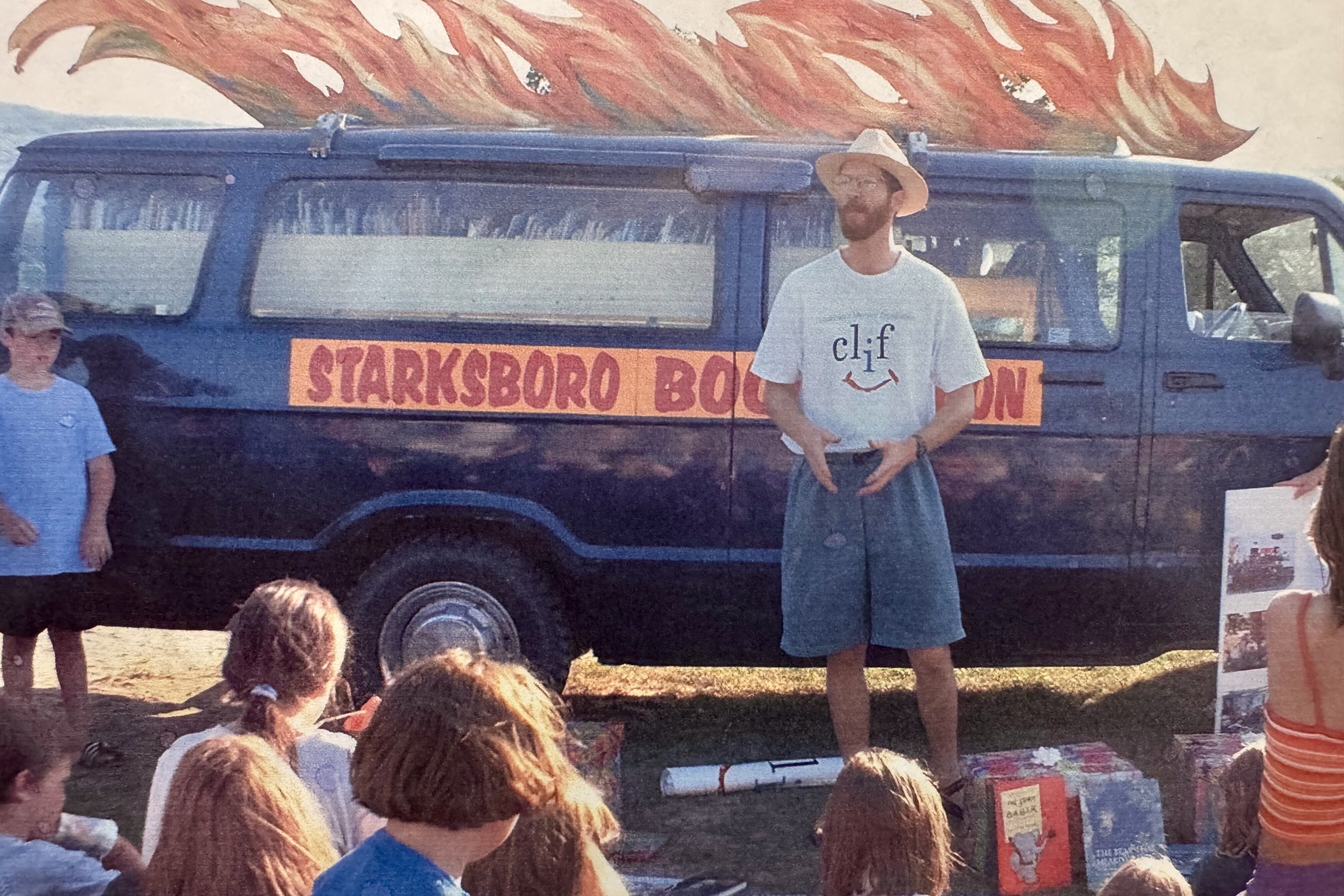 A man presents to children near a van with "Starksboro Book Bus" and flame art.