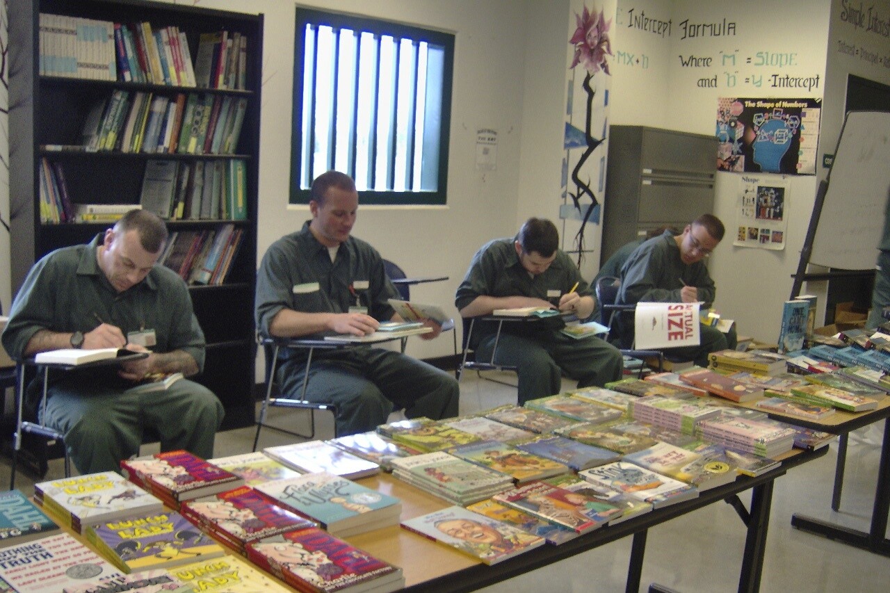 Four individuals in green uniforms write at a table with various books and educational materials.