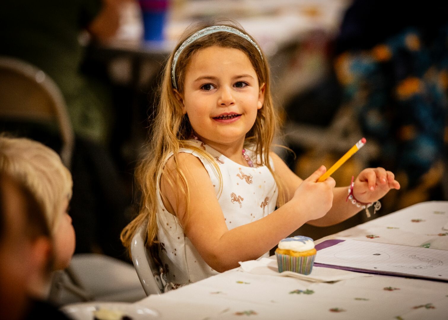 Smiling girl with long hair holds a pencil at a table with a cupcake and drawing materials.