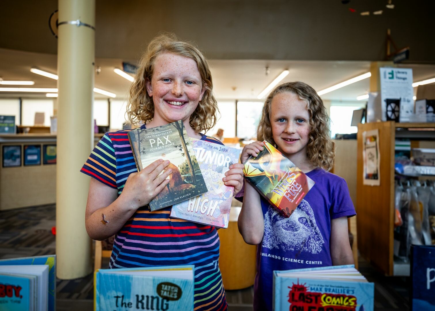 Two smiling girls hold books in a library, surrounded by shelves of children's literature.