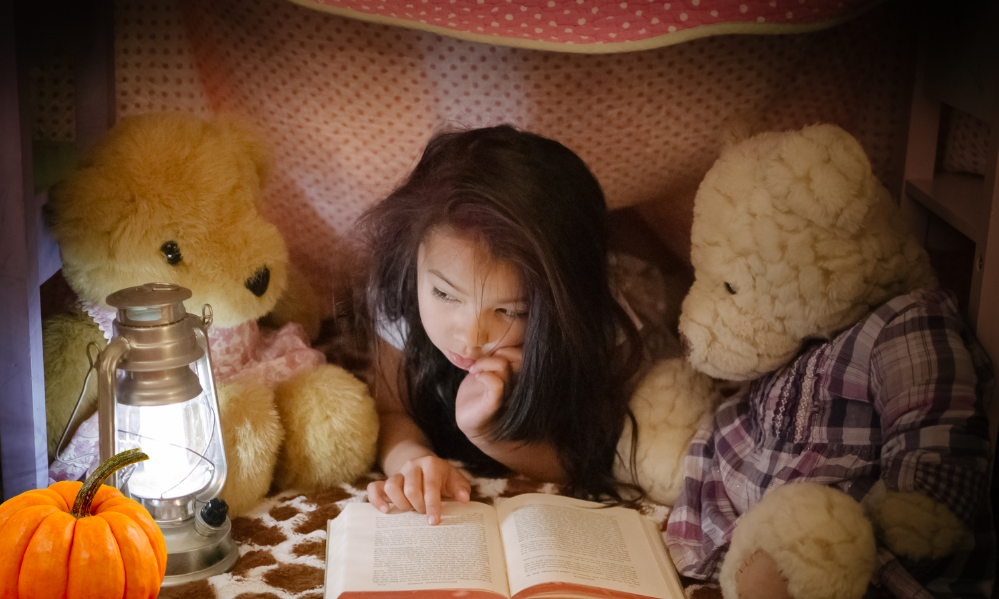 Girl lying under a blanket fort reading a book with teddy bears and a glowing lantern.