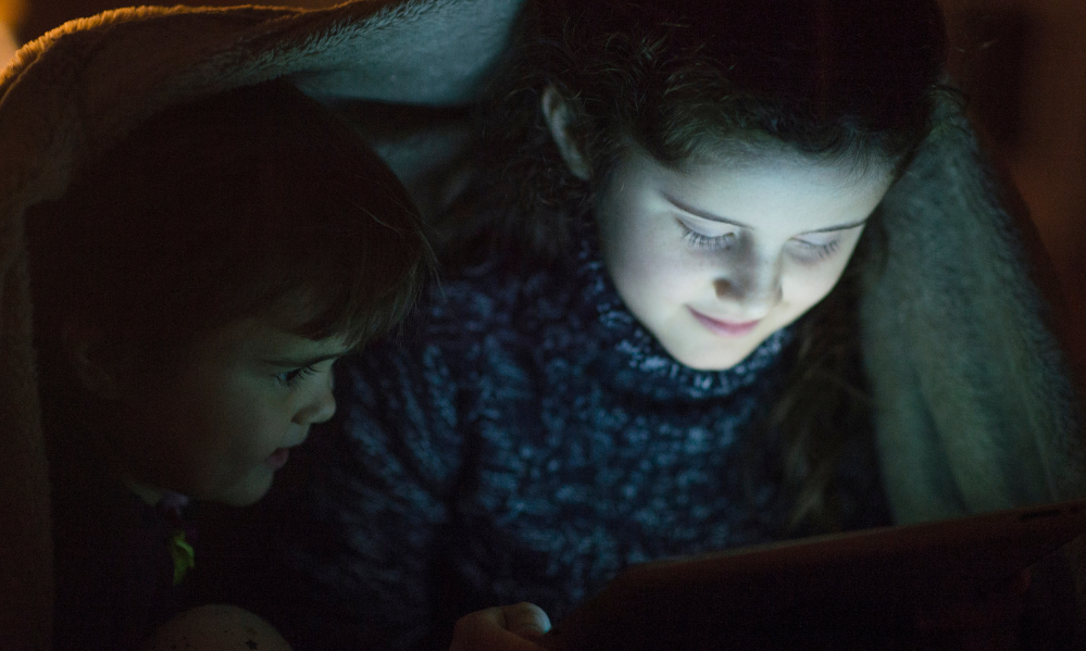 Two children sitting under a blanket looking at a glowing tablet in the dark.