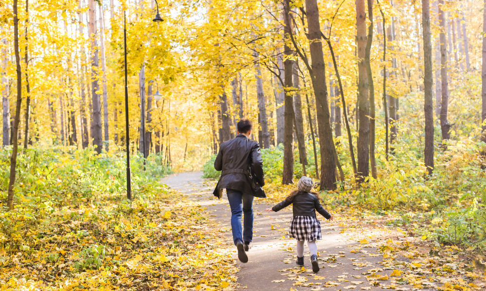 Parent and child strolling through a forest trail filled with yellow autumn leaves.