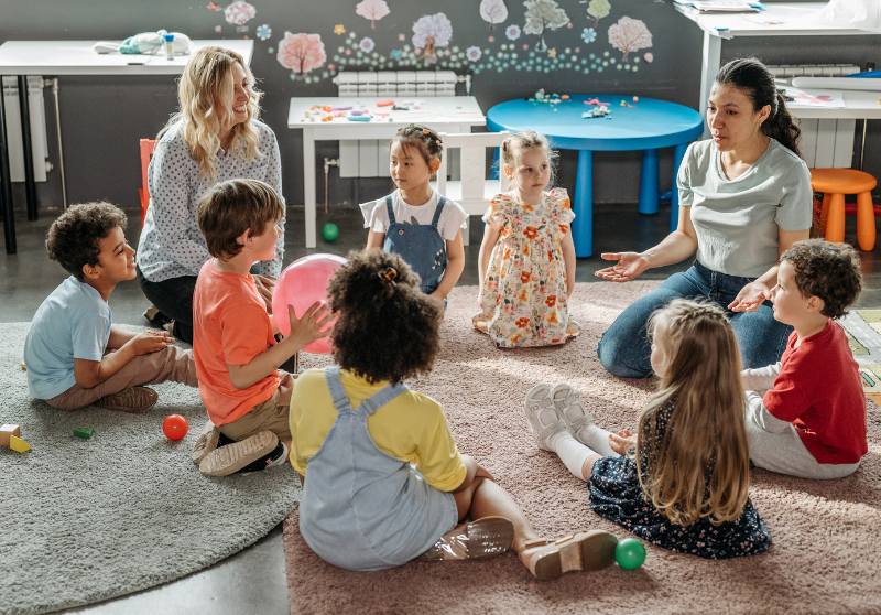 Early childhood educators guiding a small group of preschool children during circle time in a colorful classroom.