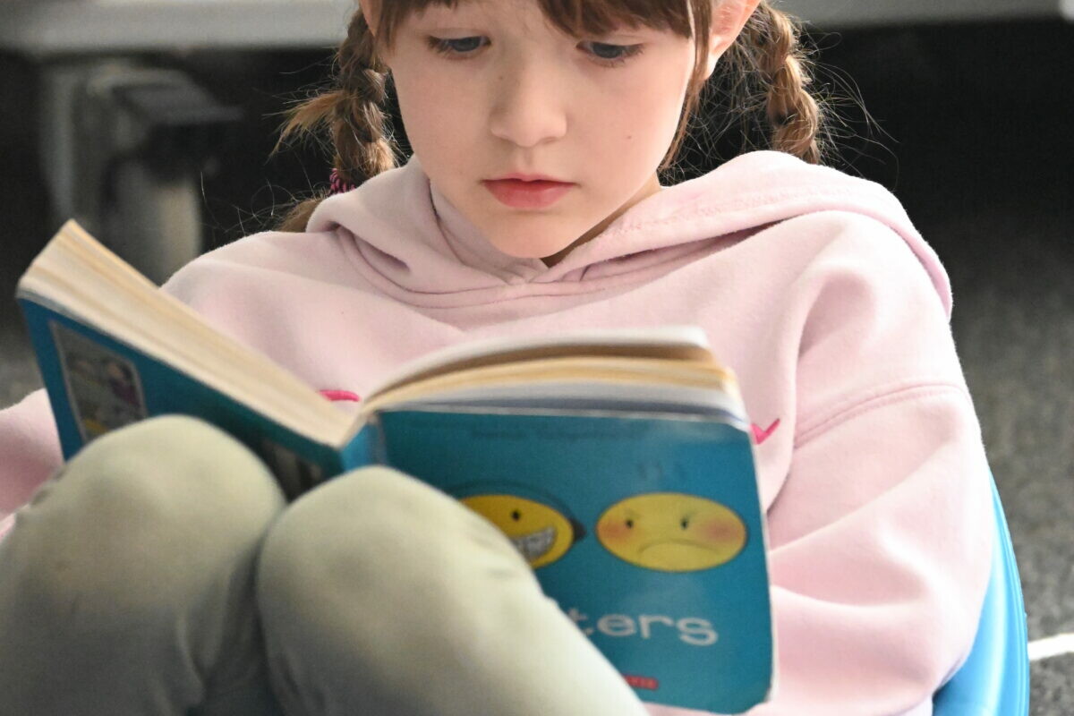 Child in pink hoodie reading a book while sitting on a blue chair.