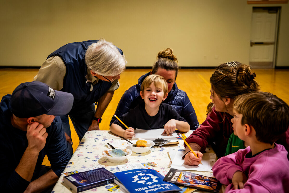 A child smiles while adults assist with art activities at a table.