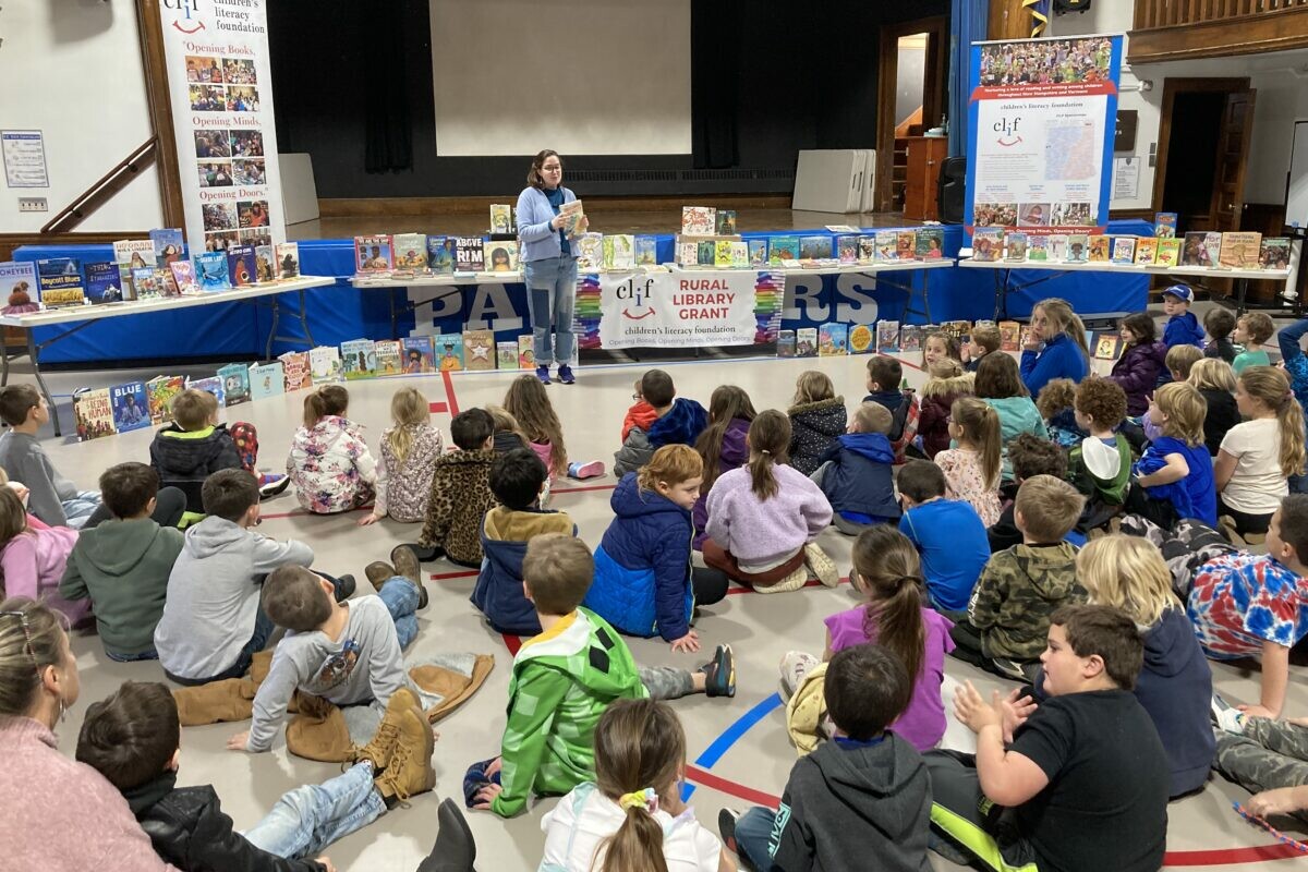 Children seated on the floor listening to a speaker in front of a table of books.