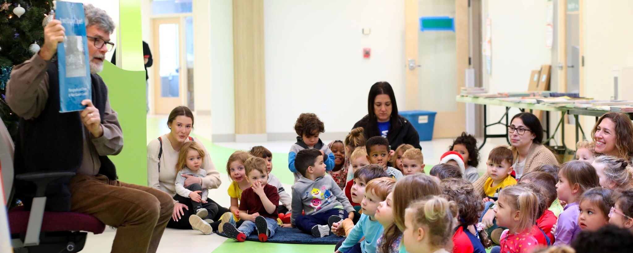 A man reads to a group of attentive children and adults in a community space.