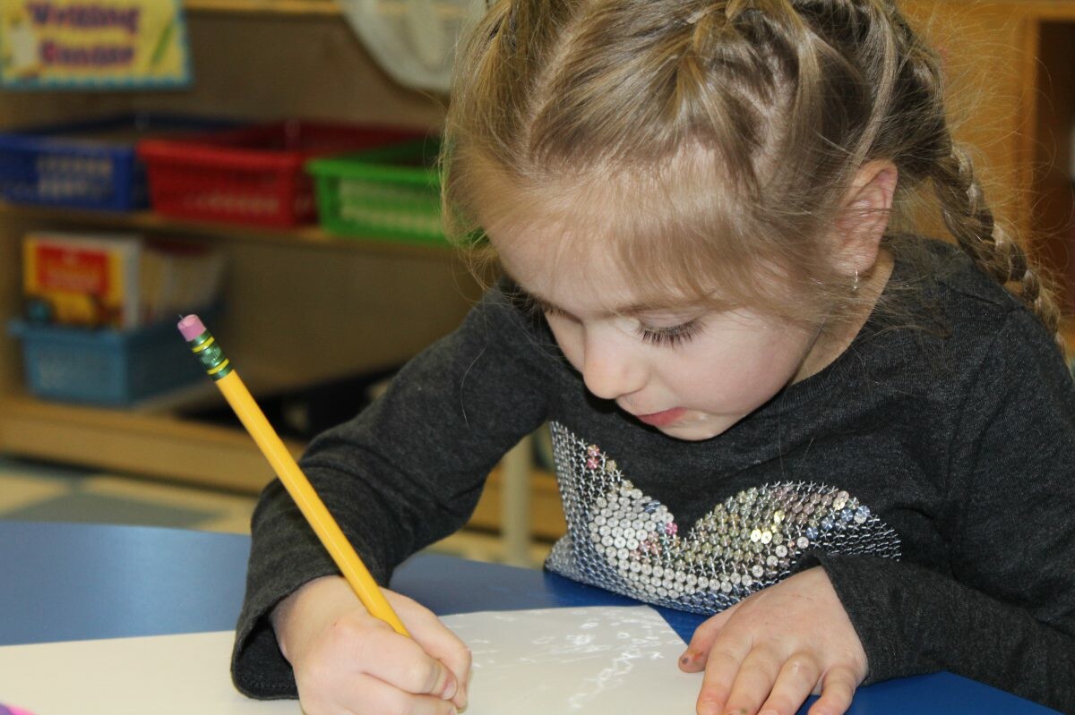 Young girl with a pencil working on art at a classroom table.