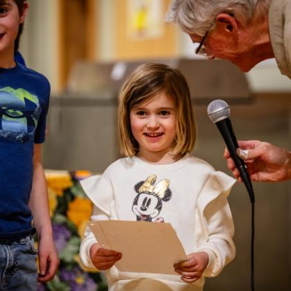 Young student holding a sheet of paper while speaking into a microphone held by an older adult.