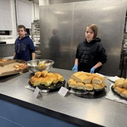 Two kids preparing trays of sandwiches at a stainless steel counter in a kitchen.