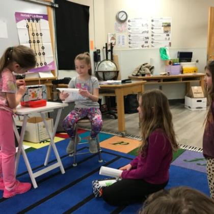 Kids participating in a classroom activity with one child standing at a small table while others watch.