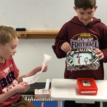 Two boys at a table examining papers and a container of small items during a classroom activity.