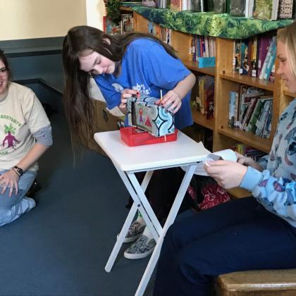 Kids gathered around a small table playing a stacking block game in a library or classroom.