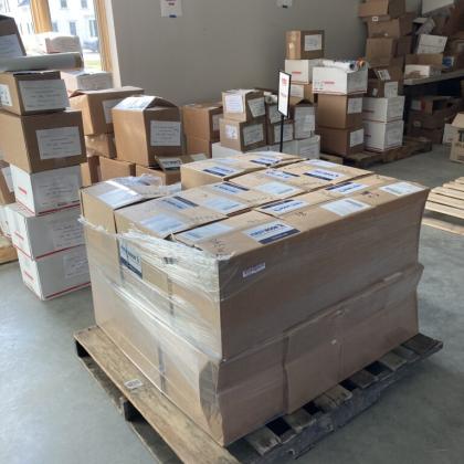 Pallet of sealed shipping boxes wrapped in plastic inside a warehouse packing area with additional labeled boxes stacked in the background.