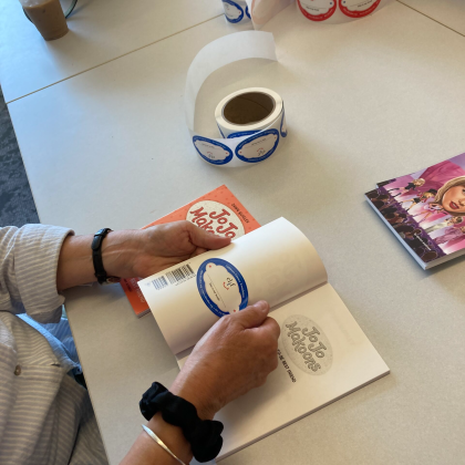 Close-up of a person placing a label inside a children’s book during packing at a book distribution workspace.