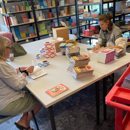 Book warehouse workspace with two people organizing and labeling stacks of children’s books surrounded by shelves of inventory.